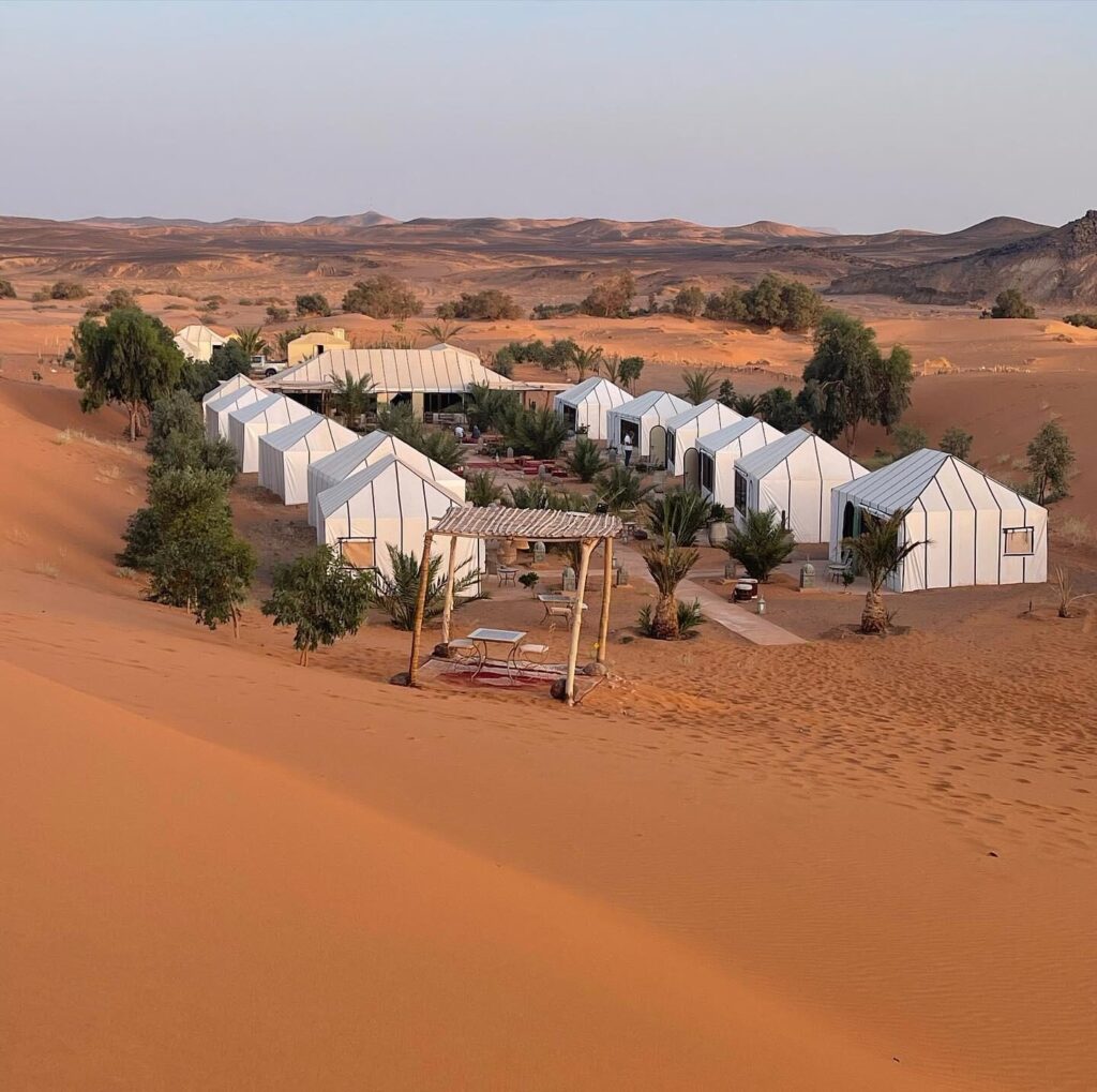 Luxury desert camp set in golden dunes with distant black desert mountains under a glowing sky.