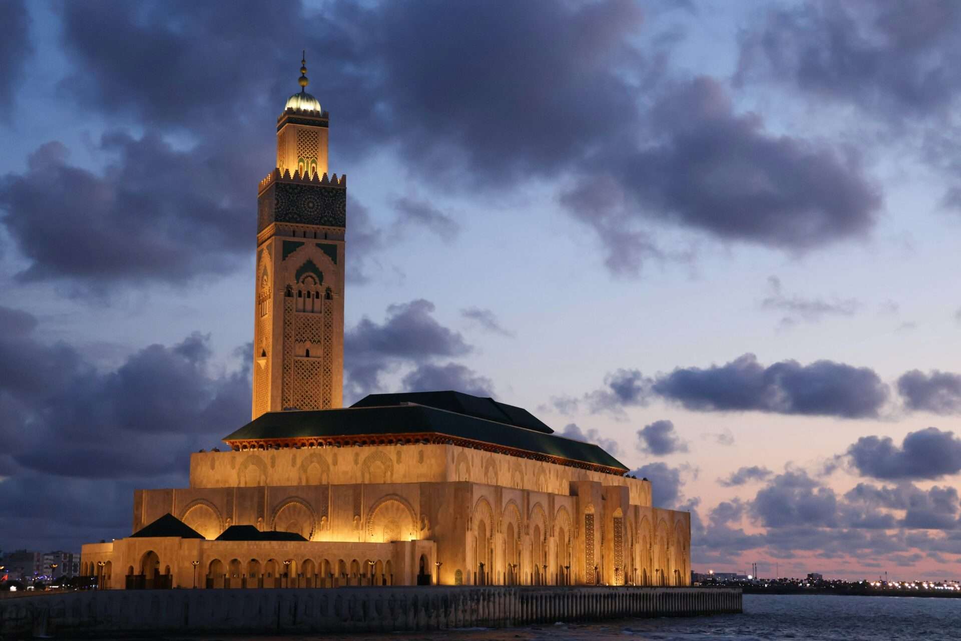 Hassan II Mosque in Casablanca beautifully illuminated by the warm colors of sunset, with its towering minaret reflecting in the Atlantic Ocean.